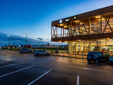 Illuminated wooden-framed building at dusk with "E.Leclerc," "Paul," and "McDonald's" signage lit up above a glass-fronted restaurant seating area, parked cars in the foreground, parked freight trucks