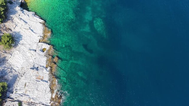 Vertical aerial view drifting along a rugged white rock coast with transparent turquoise shallows and deep blue water.