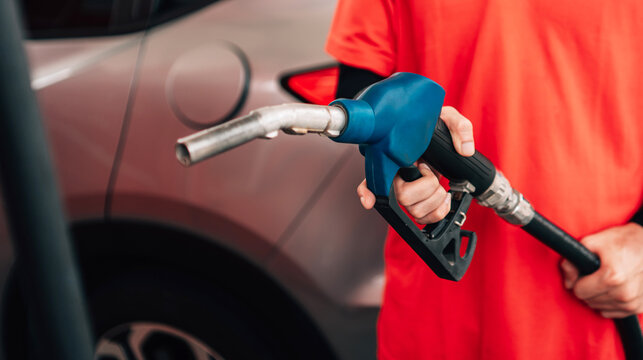 close-up, selective-focus shot at a gas station. An attendant, wearing a bright orange polo shirt and a black arm sleeve, holds out a fuel pump nozzle with a blue handle