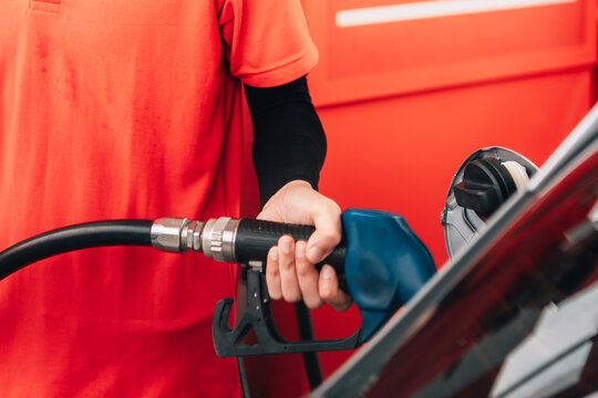 a close-up view of a person refueling a vehicle. The focus is on the individual's hand gripping a blue-handled fuel pump nozzle that is inserted into the open fuel tank of a dark car.