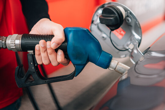 a close-up view of a person refueling a vehicle. The focus is on the individual's hand gripping a blue-handled fuel pump nozzle that is inserted into the open fuel tank of a dark car.