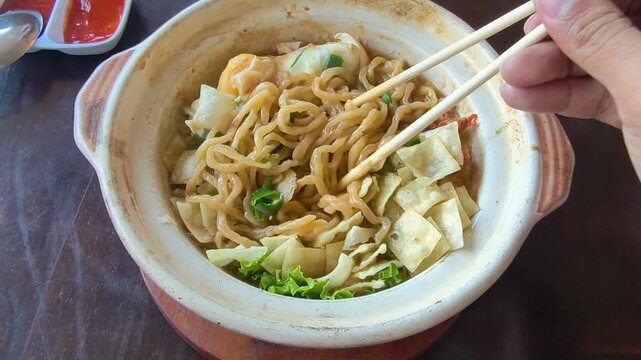 Person s hand holding chopsticks picking up delicious noodles and vegetables from a rustic earthenware bowl capturing the enjoyment of a savory meal with fresh ingredients and flavorful broth