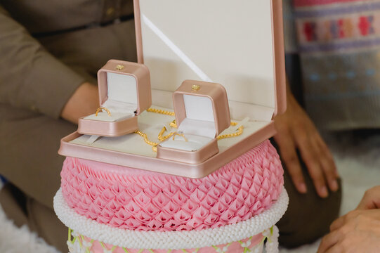 Two gold wedding rings inside pink boxes rest beside gold necklace inside traditional Thai ceremonial tray, representing dowry presented inside marriage ritual, couple sitting background indoors.