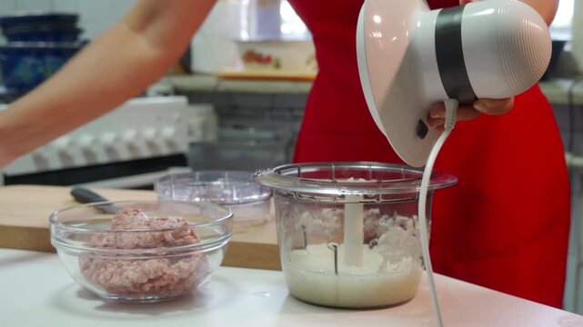 A focused woman uses a food processor next to a bowl of minced meat. She efficiently prepares a homemade meal at a kitchen counter, feeling productive and content.