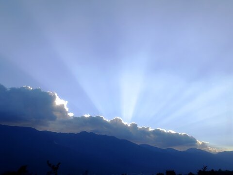 Sun rays piercing through clouds over dark mountain silhouettes, blue twilight sky landscape