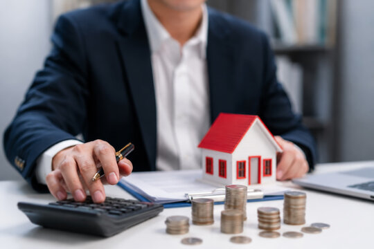 Businessman calculating home loan or mortgage investment with coin stack and house model, real estate finance planning budget and banking services. 