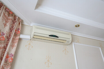 beige split-system air conditioner mounted cream wall with subtle gold fleur‑de‑lis motifs.floral-patterned curtain frames corner, while crisp white crown molding recessed ceiling light © evgeniykleymenov