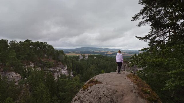Back view of a woman in a white jacket walking on a rock, enjoying the scenic view of Hruba Skala, Czech Republic