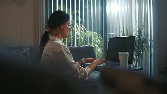 Woman working on laptop at home near window with coffee daytime focused typing and taking a short pause in a cozy environment with natural light