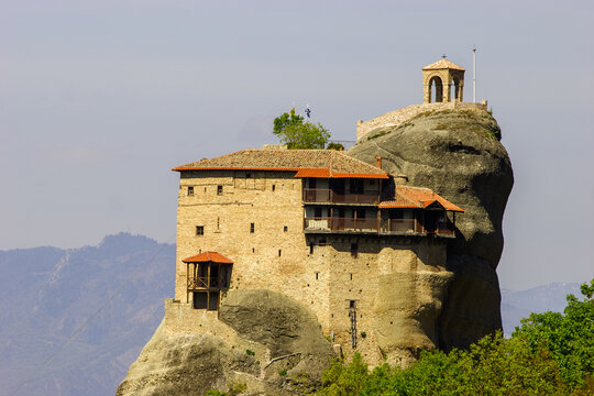 The Monastery of Great Meteoron, perched atop a rock formation in Meteora, Greece, stands against a hazy sky with distant mountains visible.