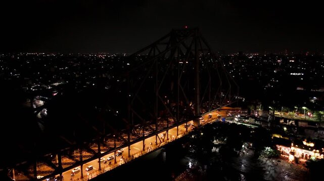 Aerial drone video of night traffic moving across illuminated Howrah Bridge in Kolkata over Hooghly River, showcasing steel cantilever structure and urban transportation.