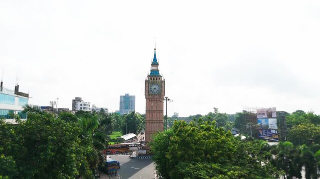 Daytime aerial drone video of Big Ben Clock Tower replica in Lake Town Kolkata, surrounded by green trees, road traffic intersection, and city landscape in West Bengal.