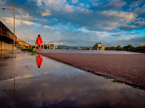 A woman in a bright red coat walking along a wet dockside promenade at golden hour, her reflection visible in a puddle, with a France T&eacute;l&eacute;visions building and Rouen's skyline in the background under a