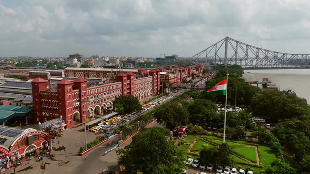 Daytime aerial drone video of Howrah Junction railway station and Howrah Bridge in Kolkata with waving Indian flag, showcasing historic red architecture, riverbank, and urban landscape.