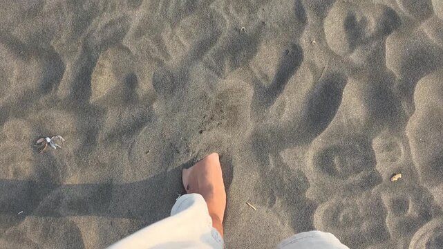 High angle view of bare feet walking on soft grey beach sand during a sunny day.