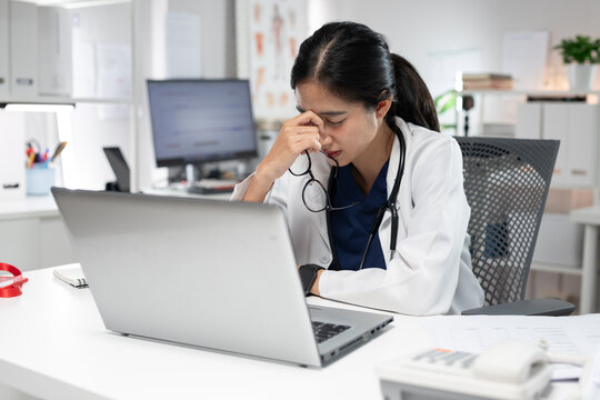 Exhausted female doctor suffering from stress and headache while working on a laptop in a medical office.Healthcare professional holding her temples after a long shift,work pressure,fatigue,