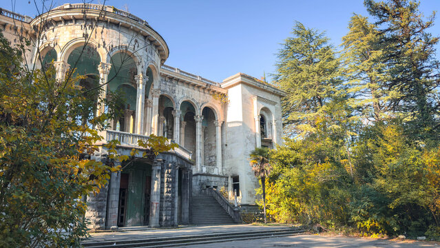 Abandoned historic sanatorium surrounded by lush trees featuring grand neoclassical colonnades, monumental arches, weathered facade, forgotten elegance in Tskaltubo, Georgia, architectural 