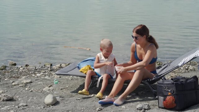 A loving mother and her small son in a life vest relax on a beach chair. They prepare to swim in the peaceful lake, where green algae grows.