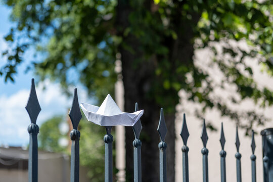 Close-up of a handmade white paper origami boat resting on the sharp metal tip of an iron fence post with beautiful natural bokeh and green trees in the background.