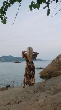 Fashion model wearing abstract print beach kimono posing on rocky tropical shoreline by calm ocean.