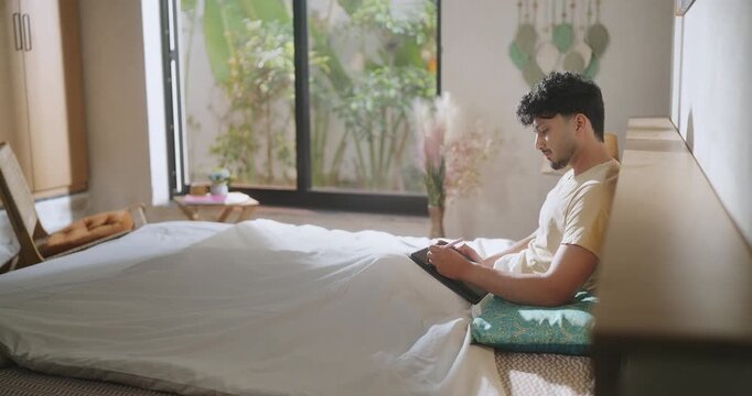 Indian young adult gen z male man sitting on bed use tab look screen do study work check email teen boy hold tablet stylus pen writing plan new notes ideas spend time take job break room home place
