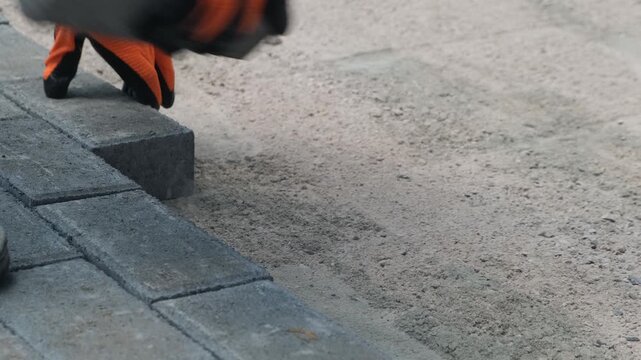 Hands of road worker in work gloves lay paving stones on foundation. Close up installation of sidewalk tiles. Urban road construction. Arrangement of sidewalks yards