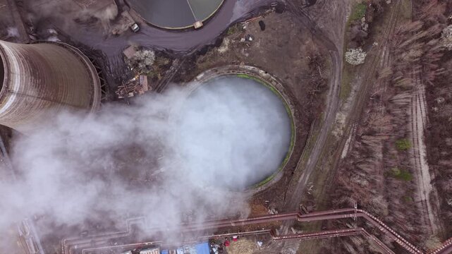 Cinematic aerial view of a thermal power station with massive cooling towers, smokestacks and industrial infrastructure. Steam rises into a dramatic cloudy sky while the nearby city highlights the rel