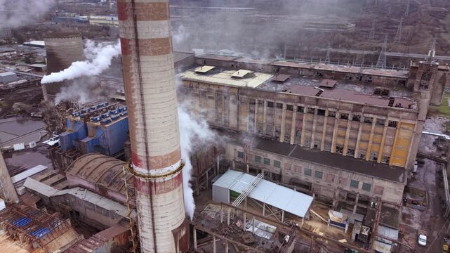 Cinematic aerial view of a thermal power station with massive cooling towers, smokestacks and industrial infrastructure. Steam rises into a dramatic cloudy sky while the nearby city highlights the rel