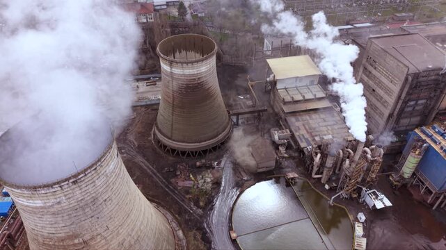 Cinematic aerial view of a thermal power station with massive cooling towers, smokestacks and industrial infrastructure. Steam rises into a dramatic cloudy sky while the nearby city highlights the rel
