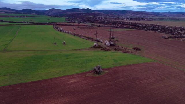 Aerial view of broad agricultural fields with green crops and freshly cultivated soil crossed by high-voltage electricity pylons and power lines, with rolling hills and rural settlements in the distan