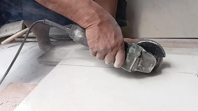 A man's hands cutting a ceramic floor tile with an angle grinder during indoor renovation. Construction worker trimming tiles with precision for flooring installation.