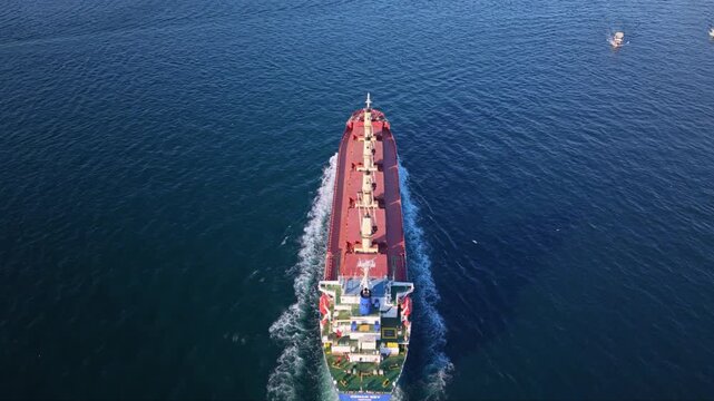 Aerial approach above a cargo ship sailing through blue water, following its white bridge and row of deck cranes toward the bow and distant boats.