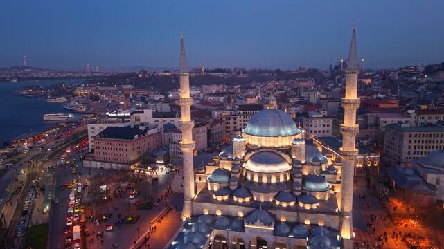 Cinematic aerial view of the New Mosque (Yeni Cami) illuminated during blue hour overlooking Emin&ouml;n&uuml; Square in the heart of Istanbul, Turkey. Historic Ottoman architecture, lively streets and the vibr