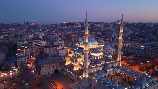 Cinematic aerial view of the New Mosque (Yeni Cami) illuminated during blue hour overlooking Emin&ouml;n&uuml; Square in the heart of Istanbul, Turkey. Historic Ottoman architecture, lively streets and the vibr