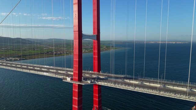 Close aerial pass beside a red suspension bridge tower and white vertical cables, with the roadway crossing deep blue water above a small passing vessel.