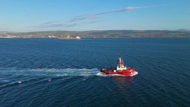High-resolution aerial footage of a harbor tugboat navigating open water near commercial shipping lanes with a cargo ship in the distance and coastal hills on the horizon. Ideal for maritime transport