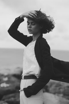 Moody black and white scene of a wind swept girl wearing a blazer and white dress by the sea, hand in hair, calm expression, movement and ocean breeze captured with minimalist styling