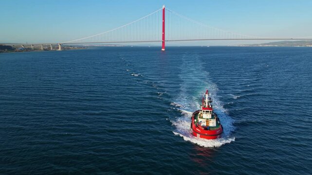 High-resolution aerial footage of a harbor tugboat navigating open water near commercial shipping lanes with a cargo ship in the distance and coastal hills on the horizon. Ideal for maritime transport