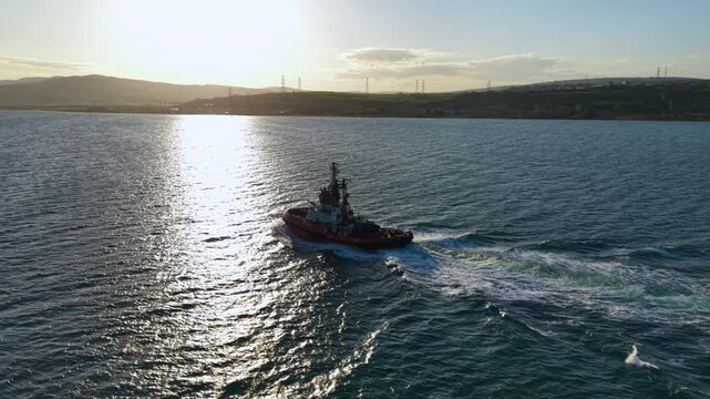 High-resolution aerial footage of a harbor tugboat navigating open water near commercial shipping lanes with a cargo ship in the distance and coastal hills on the horizon. Ideal for maritime transport