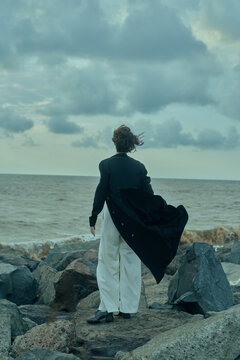 Figure standing on rocky shore in cape and tailored attire, facing the sea as wind sweeps the cape, creating a dramatic coastal moment, fusion of formalwear and rugged landscape, moody sky, expansive