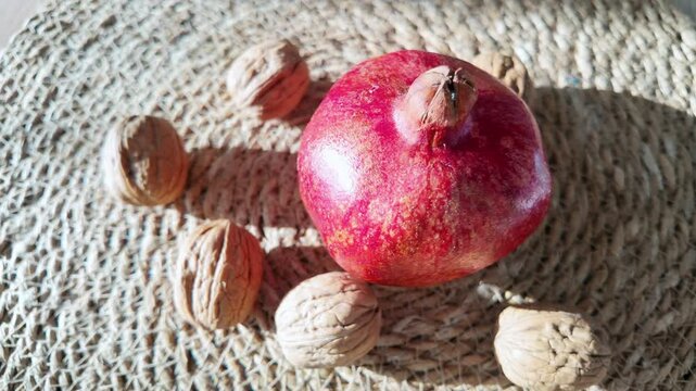 Fresh ripe pomegranate fruit surrounded by whole walnuts on woven placemat in natural sunlight.