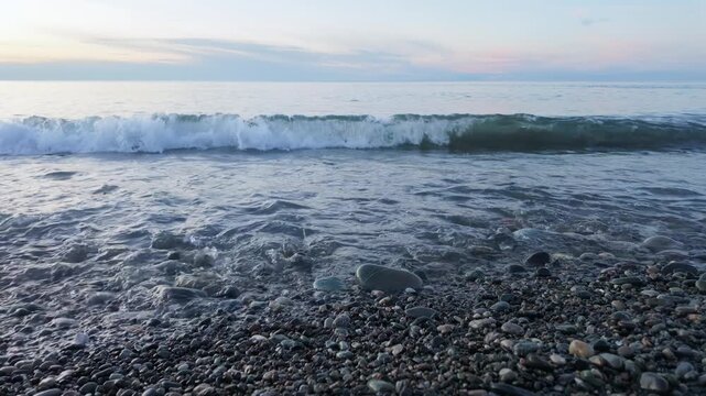 Gentle ocean wave rolling and breaking onto pebble beach shoreline at calm dusk with soft sky.