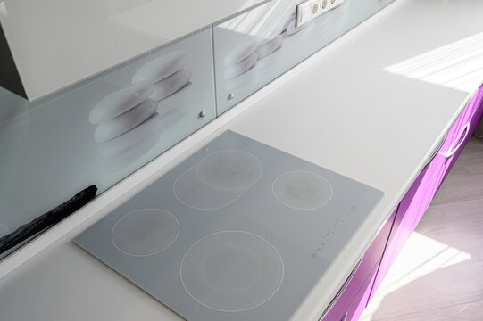 modern kitchen features a sleek gray glass‑ceramic four‑burner cooktop on a white countertop, paired with purple lower cabinets and a minimalist glass backsplash with a subtle plate motif