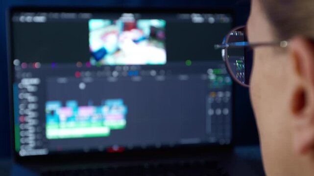 Soft focus shot of woman viewing blurred laptop screen displaying video editing software timeline. Indistinct interface reflected in glasses of female editor working on digital media production.