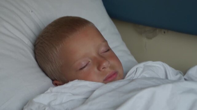 A peaceful young boy sleeps deeply tucked under a white blanket. He rests calmly on a soft pillow inside a quiet train compartment during a long family journey.