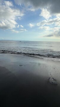Dark sand beach with gentle ocean waves and cloudy blue sky