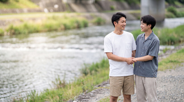 夏の川沿いの道で手を繋ぎ優しく微笑み合う男性二人 ｜ 爽やかな風