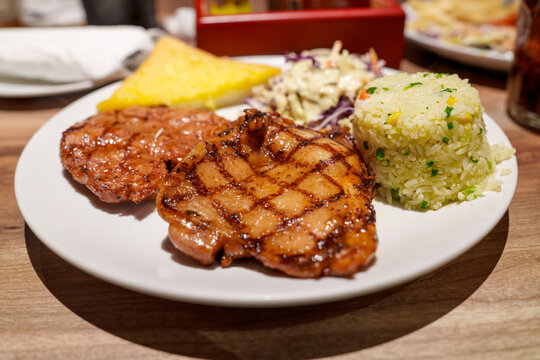 Grilled chicken fillet with grill marks served on a white plate with fried rice, coleslaw, and a side of garlic bread. Delicious restaurant meal served on a wooden table. Healthy protein dish.