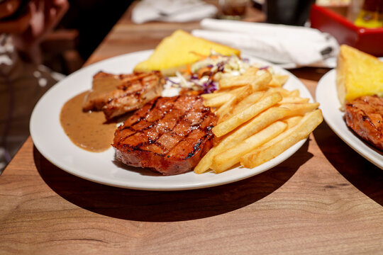 Grilled pork steak and chicken fillet served with golden french fries, gravy, salad and garlic bread on a white plate in a restaurant. Gourmet meat dinner meal on a wooden table. Close up view.