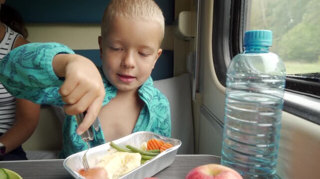 A happy boy holds a sausage on a fork while eating lunch inside a moving train. This cozy travel meal brings a cheerful and comforting feeling to his journey.